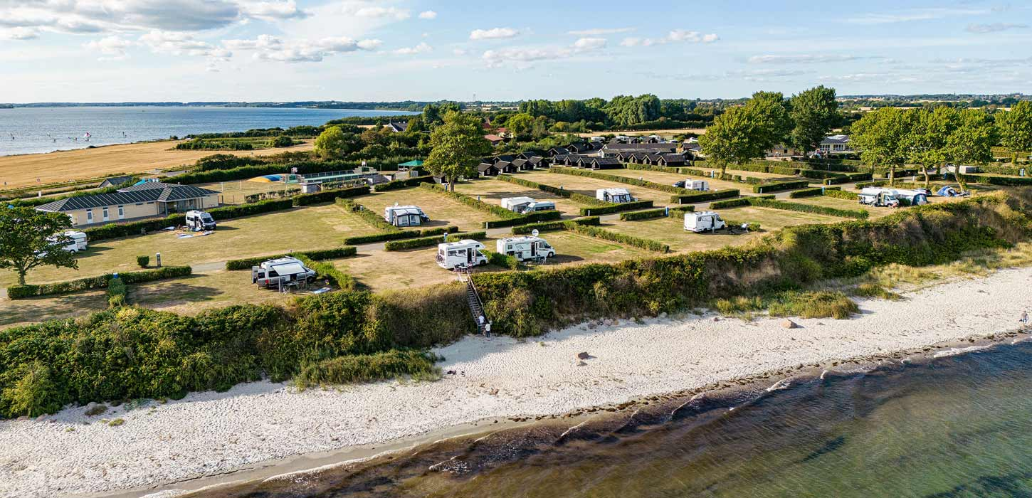 Drejby Strandcamping — luftfoto af campingplads med grønne plæner, campingvogne, hytter og faciliteter langs en hvid sandstrand med klart vand og udsigt over havet