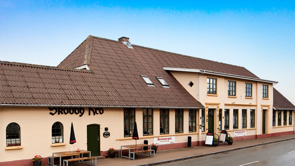 Skovby Kro — klassisk dansk kro med cremefarvet facade, brunt tegltag, grønne døre og udendørs borde med parasoller langs gaden