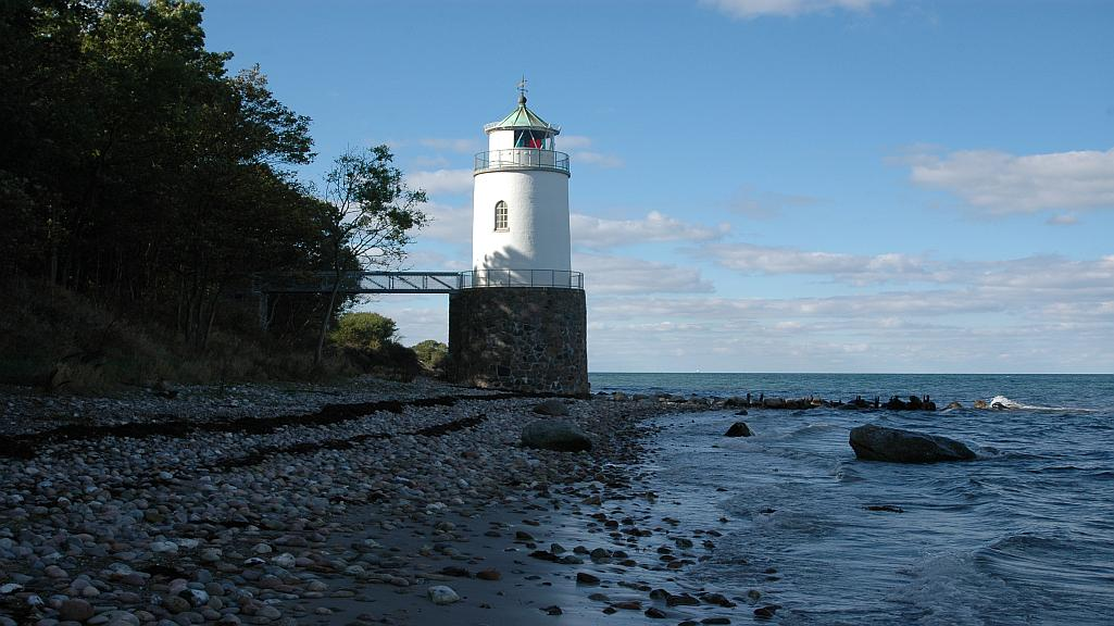 Alsstien — hvidt fyrtårn på stenede strand ved det klare blå hav med skov og gangbro langs kysten under sommerhimmel