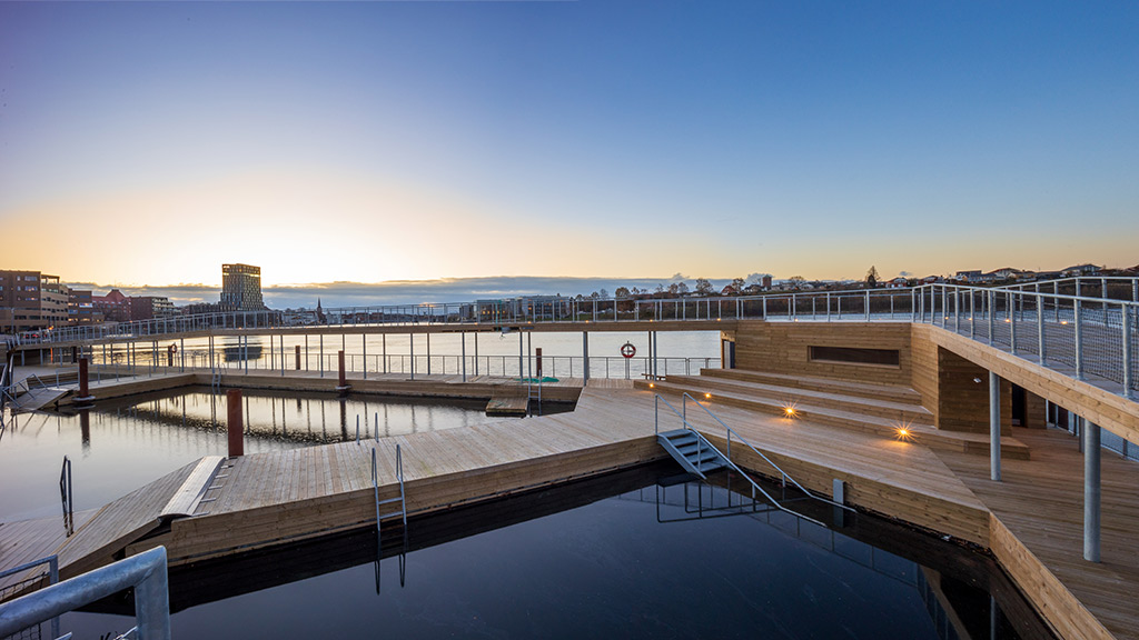 Sønderborg Havnebad — moderne træbrygge med bassiner og trapper ned i vandet ved solnedgang, med byens tårne og havn i baggrunden