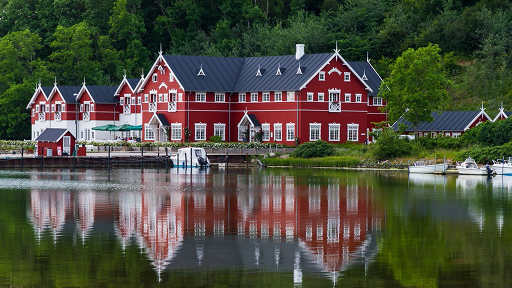 Dyvig Badehotel — karakteristisk rødt hotel med hvidt bindingsværk og mørkt tag, spejlet i det stille vand med både ved broen og grønne træer i baggrunden