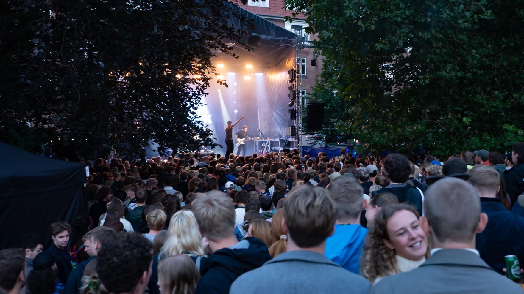 Kultur året rundt — udendørskoncert med stor menneskemængde foran en scene med gule lys og træer i aftenlys, musiker på scenen med armene i vejret