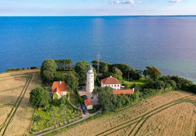 Kegnæs Fyr — luftfoto af det hvide fyrtårn med rødt tag, omgivet af grønne træer og marker, med Østersøens klare blå vand så langt øjet rækker