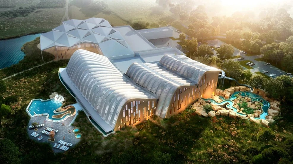 Center Parcs Nordborg Resort — luftfoto af stort feriecenter med bølgede glastage, udendørs poolområde med rutsjebaner, vandland og grønne omgivelser mod en solnedgangshimmel