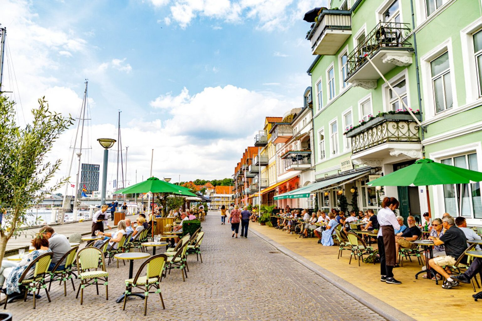 Sønderborg Havnepromenade — livlig gade med farverige bygninger, grønne parasoller, gæster der spiser udendørs på caféer og restauranter med sejlbåde i havnen i baggrunden