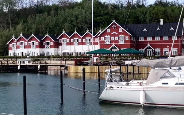 Dyvig Badehotel, det røde hotel med hvide vinduer langs Dyvig-vigen, lystbåde på vandet foran og skov på den anden side