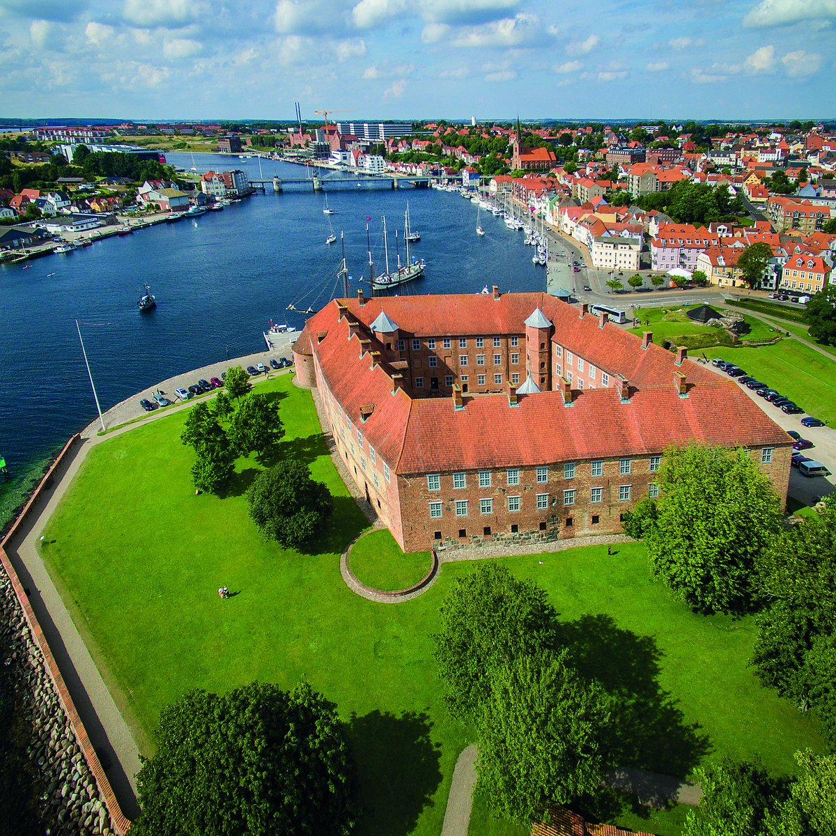 Luftfoto af Sønderborg Slot med rødt tegltag, omgivet af grønne volde med Als Sund, havn og by i baggrunden
