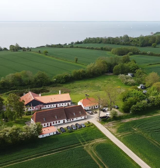 Luftfoto af Als Kloster — hvide bygninger med røde og orange tegltage, omgivet af grønne marker og med havet i baggrunden