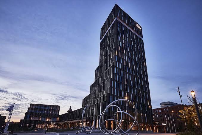 Hotel Alsik — moderne højt hotel i mørke facadepaneler mod en skumringshimmel, med kunstværker på pladsen foran og havnen i baggrunden