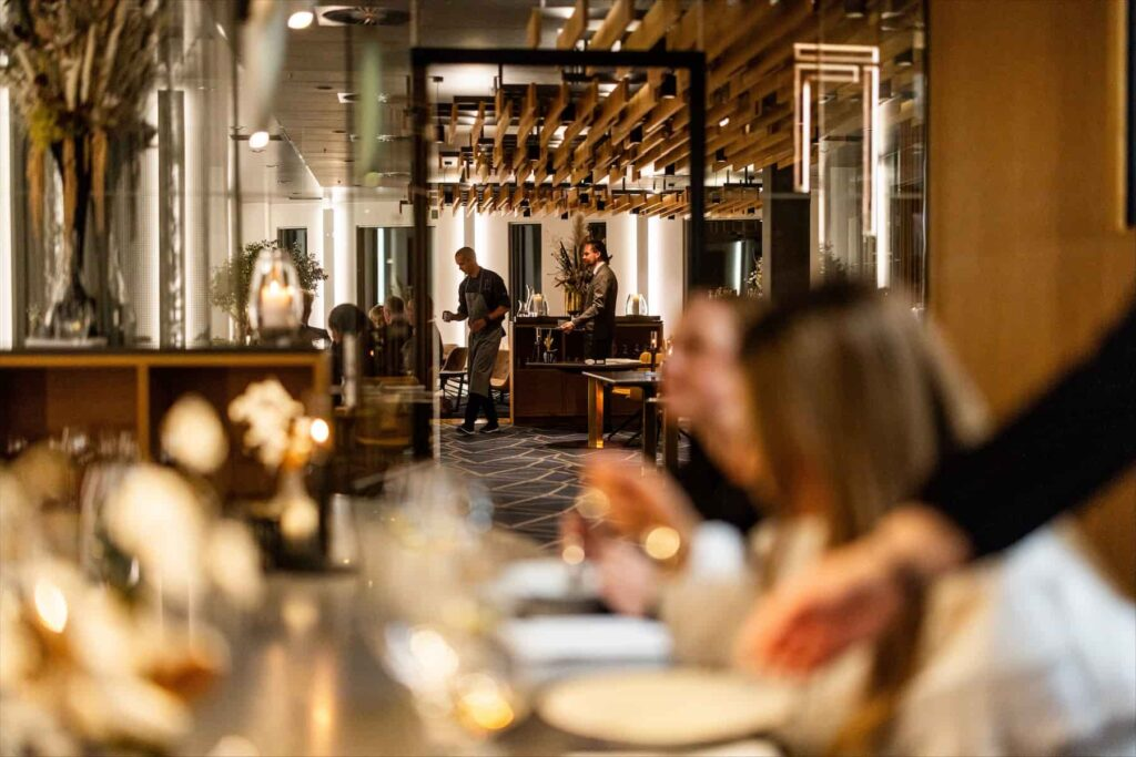 Restaurant Syttende — elegant restaurantinteriør med træloftsdetaljer, blødt lys, gæster ved dækkede borde og køkkenpersonale i baggrunden