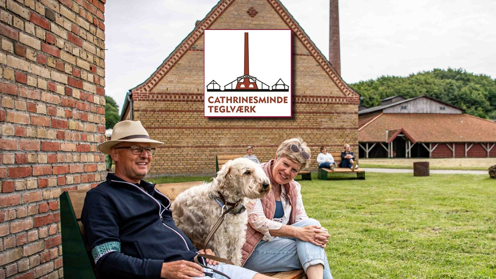 Cathrinesminde Teglværk — et par med en stor, krøllet hund sidder på en bænk foran et gammelt gule murstensbygning med rødt tegltag og industriskorsten, grøn græsplæne og træer i baggrunden